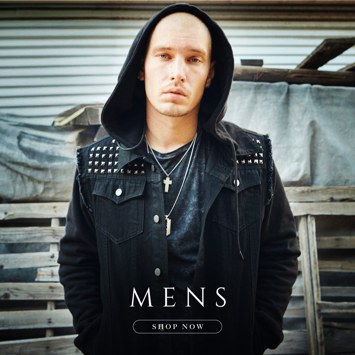 Man wearing a black jacket with studs and a hood, standing in front of a rustic background with 'MENS SHOP NOW' text.