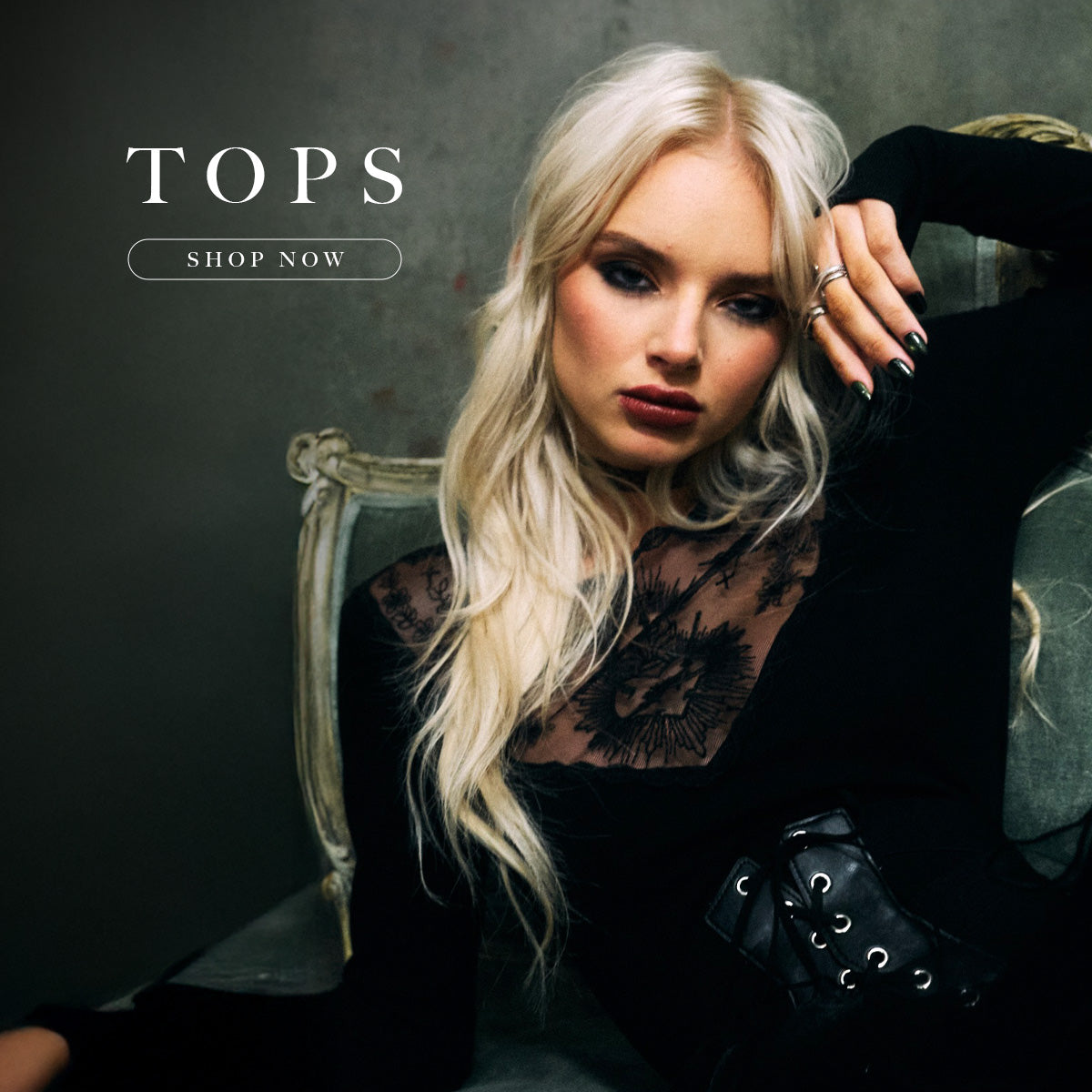 Woman with blonde hair wearing a black top with lace details, sitting on a chair against a dark background. 'TOPS SHOP NOW' text is displayed.